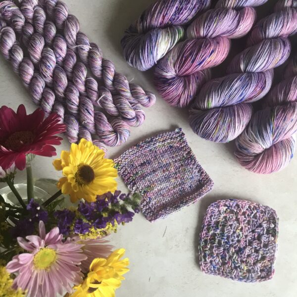 a posy of bright yellow and pink flowers is on the bottom left corner of an off white, plaster background. Knit and crochet swatches are in the centre with full skiens to the top right and minis to the top left. The yarn is a mix of soft pinks and purples, with flashes of white and lots of pink, blue, green and yellow speckles.