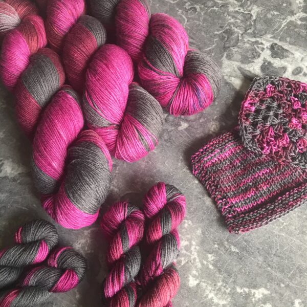 on a grey marble background are three full sized skiens at the top left and 4 mini skeins in the bottom left and centre. knit and crochet swatches are to the centre right. the yarn is a regular repeating variegated colour way shifting from charcoal grey into deep red and raspberry pink.