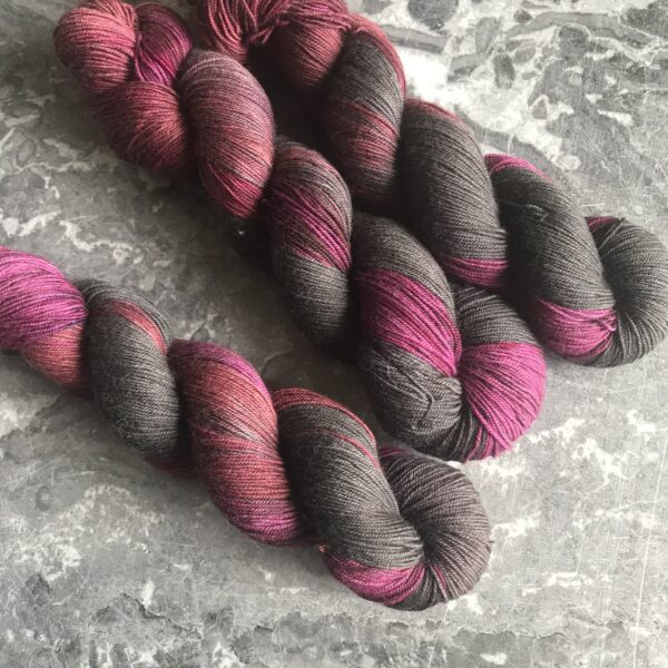 on a grey marble background are three twisted skeins, placed an an angle from the top left corner. the yarn has a slight sheen to it, and rich tones of black, red and rapsberry pink that shift from one to other along the variegated skeins.