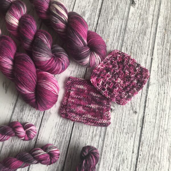 on a rustic, white wooden background are three full and three mini skeins as well as knit and crochet swatches. The yarn is deep raspberry pinks with a hint of purple and white, with lots of pink and black speckles all over.