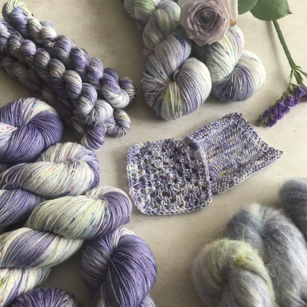 in the centre of an off white, plaster background are knit and crochet swatches, surrounded by clusters of skeins in varying weights, as well as a soft lilac rose and deeper purple flowers. The yarn is a soft lavender and white mix, with tonal areas and speckles in pink, blue and green.