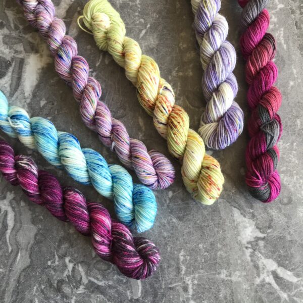 on a grey marble background are 6 mini skeins, all coming from the top left corner and slightly spaced out. The outer two minis are darker pink and black tones (one varigated, one heavily speckled). Between them are softer shades of pale blue, lavender, pale green and lilac pink, each speckled with soft tones.